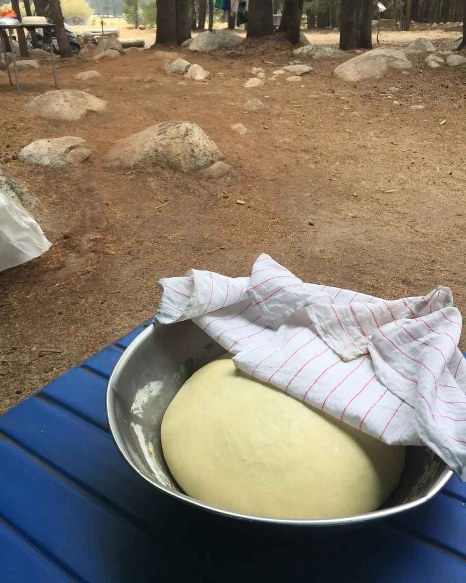 Fresh pizza dough proofing in a covered metal bowl on a camp table