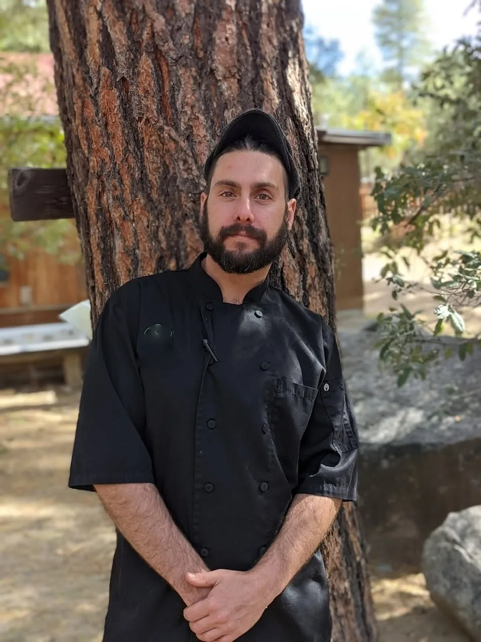 Jason Blazer in a black chef coat outdoors beside a pine tree