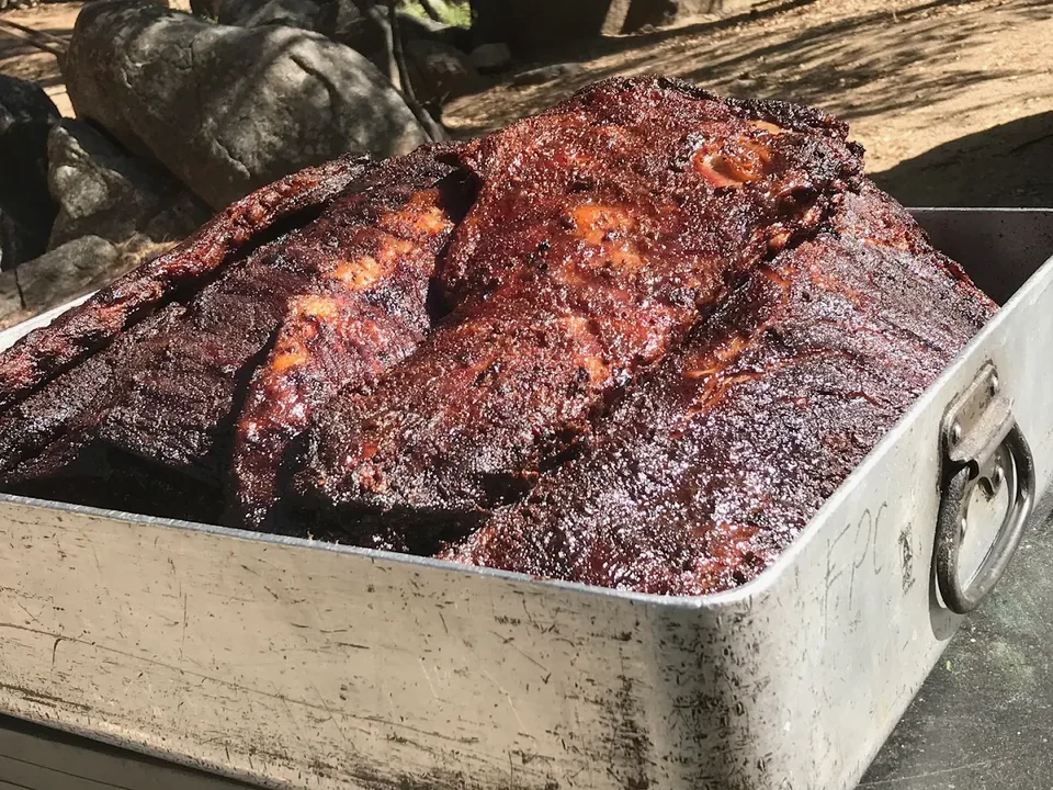Darkly glazed cooked meat piled in a metal roasting pan outdoors